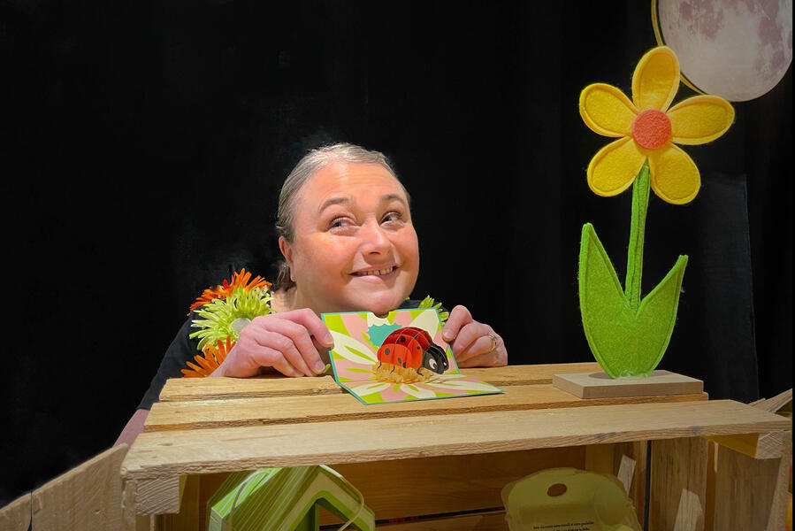 J'me cache ! La chanteuse Aurore, avec un grand sourire tient sa coccinelle et son livre à côté de sa fleur au clair de lune.
